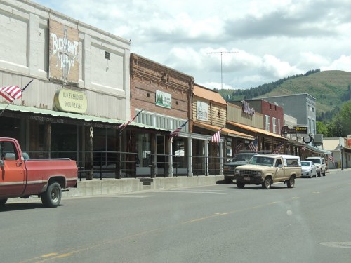 A quaint street lined with historic buildings and shops, featuring American flags and parked trucks under a cloudy sky.