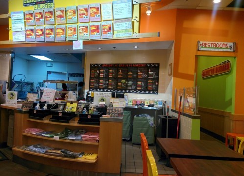 Interior of a fast-food restaurant with a colorful menu board, counter, and restrooms sign visible.
