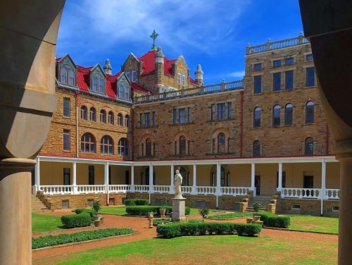 Historic stone building with red roofs, surrounded by a green courtyard and a statue, under a blue sky.