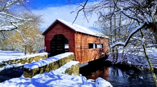 A wooden covered bridge over a stream, surrounded by snow-covered trees and a clear blue sky.