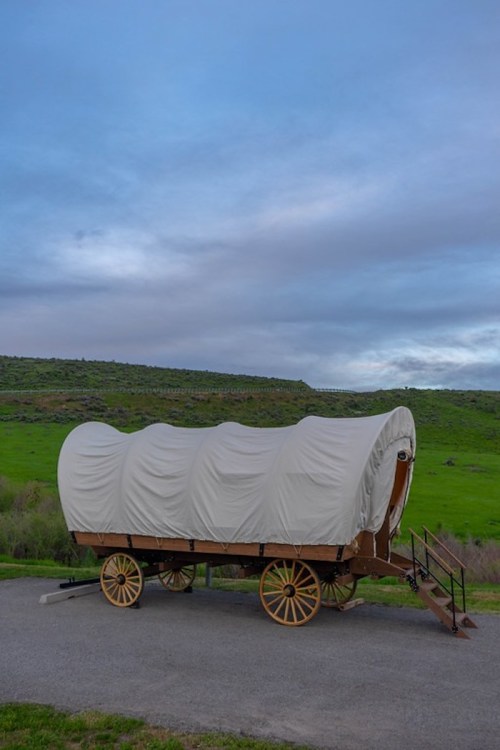 A covered wagon on a gravel path, set against a green hillside under a cloudy sky.