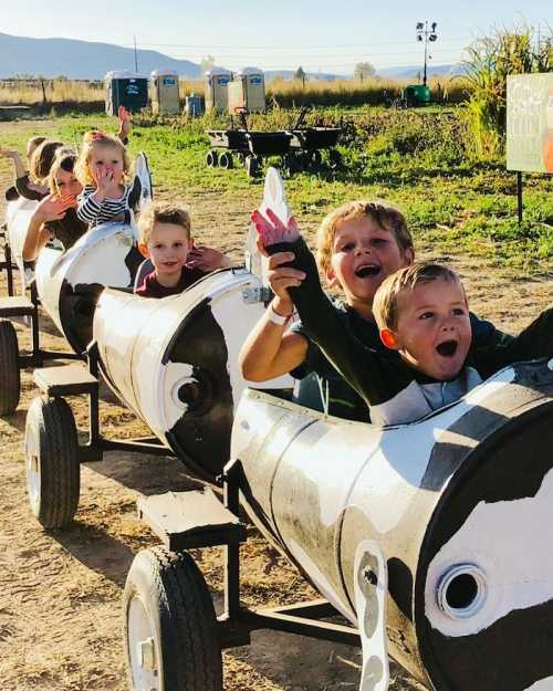 Children joyfully ride a train made of painted barrels, waving their hands in a sunny outdoor setting.