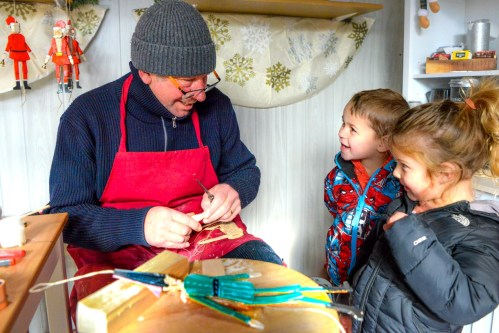 A craftsman carves a wooden figure while two smiling children watch in a cozy workshop.