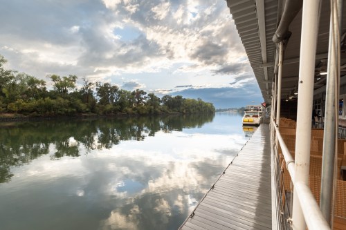 A calm river scene with reflections of clouds and trees, alongside a wooden dock and a boat.