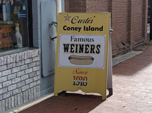 A yellow sandwich board sign for Curtis' Coney Island, advertising "Famous Weiners" since 1918.
