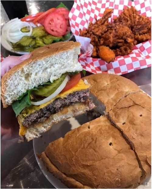 A hand holding a burger with lettuce, tomato, pickles, and cheese, with fried chicken and sides in the background.