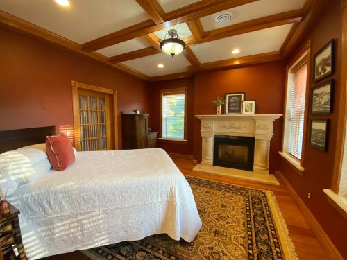 Cozy bedroom with a white bed, fireplace, and rich brown walls, featuring wooden beams and a decorative rug.