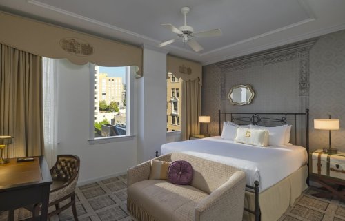 A stylish hotel room featuring a large bed, seating area, desk, and windows with city views.
