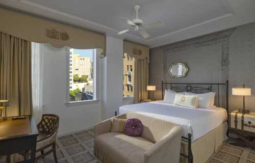 A stylish hotel room featuring a large bed, seating area, desk, and windows with city views.