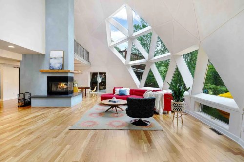 Modern living room with large windows, a red sofa, a fireplace, and wooden floors, surrounded by greenery.