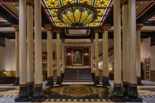 Elegant lobby with tall columns, a stained glass ceiling, and a grand staircase leading to an upper level.