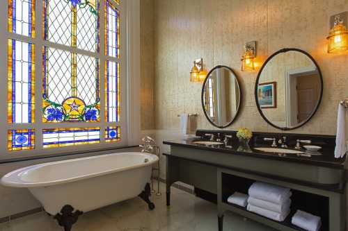 Elegant bathroom featuring a vintage bathtub, dual mirrors, and a colorful stained glass window.