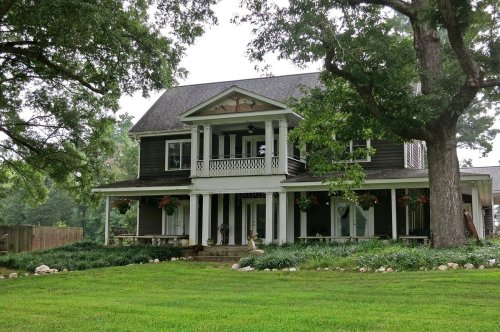 A large, two-story house with a porch, surrounded by greenery and trees, set in a spacious yard.
