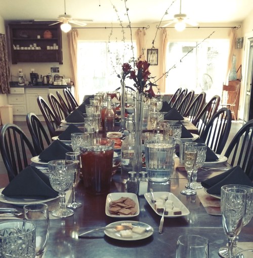 A long dining table set for a meal, adorned with glasses, plates, and a floral centerpiece in a bright room.