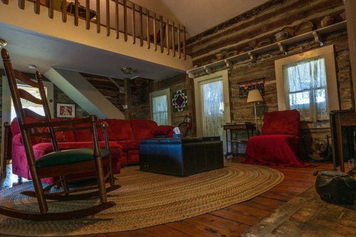 Cozy rustic living room with a red couch, rocking chair, and wooden accents, featuring a braided rug and large windows.