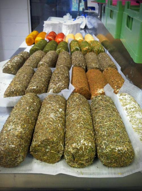 A variety of colorful, herb-covered logs of cheese arranged on a table, with some wrapped in parchment paper.
