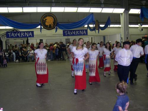 A group of dancers in traditional attire perform in a festive hall decorated with blue banners and a crowd watching.