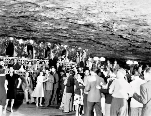 A lively dance scene in a cave, with people in vintage attire enjoying music and socializing under dim lights.