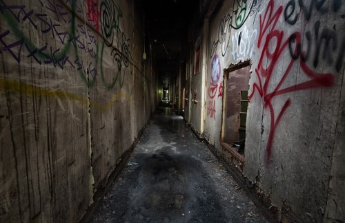 A dimly lit, abandoned hallway with graffiti-covered walls and a worn, dirty floor.
