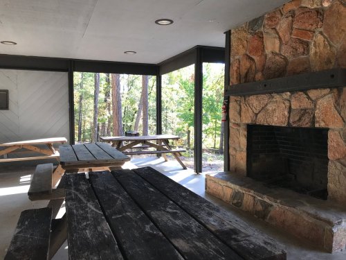 Interior of a rustic shelter with wooden picnic tables and a stone fireplace, surrounded by trees.