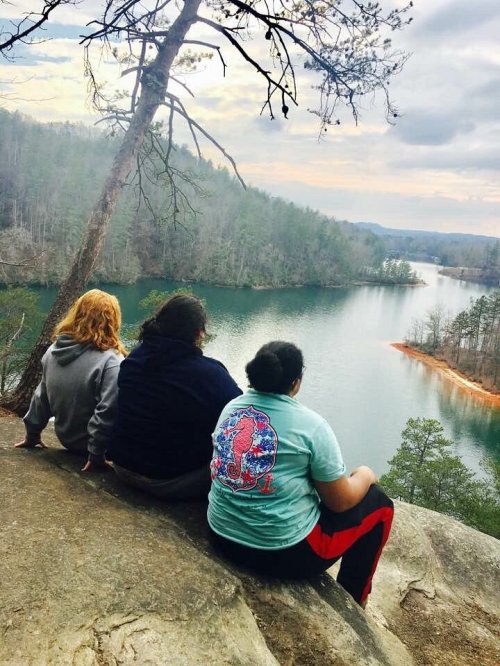 Three people sit on a rock overlooking a serene lake surrounded by trees and hills under a cloudy sky.
