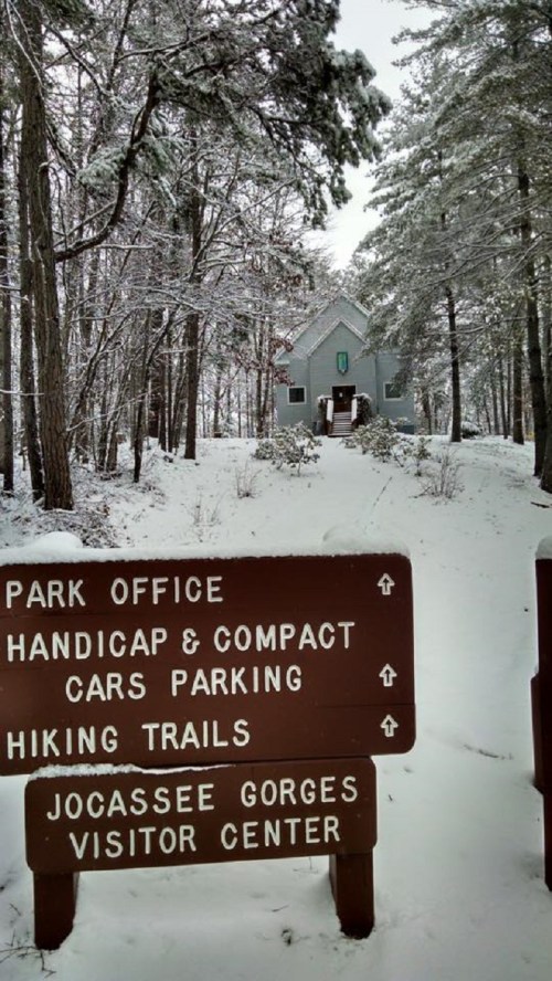 Snow-covered landscape with a park office and visitor center sign, surrounded by tall trees.