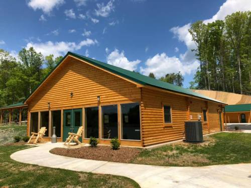 A wooden cabin with a green roof, large windows, and a patio, surrounded by trees and blue skies.