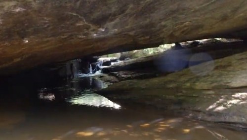 A serene view of a rocky cave with a small waterfall and calm water reflecting the surrounding nature.