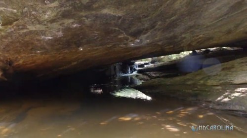 A serene view of a rocky creek with water flowing under a large stone overhang. Sunlight reflects on the water's surface.
