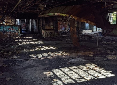 Abandoned interior with graffiti, debris, and sunlight casting shadows on the floor through broken windows.