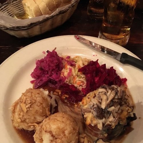 A plate of food featuring meat, mashed potatoes, red cabbage, coleslaw, and beets, with bread and drinks in the background.