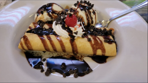 A dessert plate featuring a chocolate-drizzled crepe, topped with whipped cream, chocolate chips, and two scoops of ice cream.
