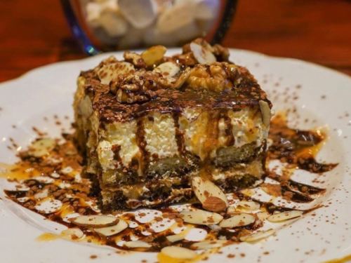 A slice of layered dessert topped with chocolate drizzle, nuts, and cocoa powder, served on a white plate.