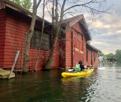 A person in a yellow kayak paddles past a partially submerged red house surrounded by water and trees.