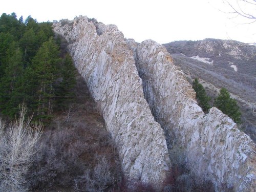 A steep, rocky cliffside with jagged formations, surrounded by sparse trees and a mountainous landscape.