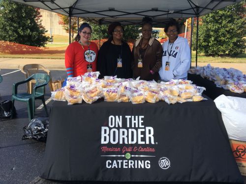 Four women stand behind a table with food, featuring an "On the Border" catering banner, under a tent.