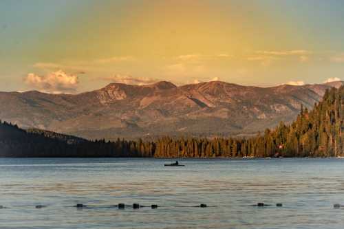 A serene lake surrounded by mountains and trees, with a lone canoeist paddling in the calm water at sunset.