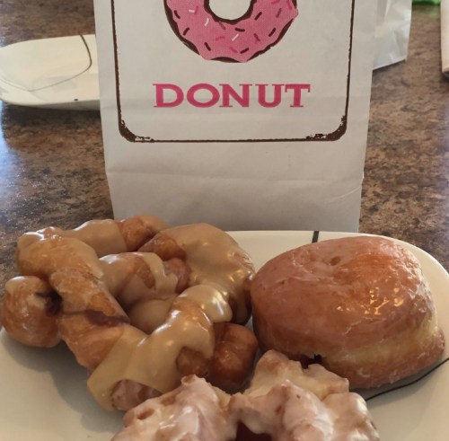 A plate with three donuts: a glazed round donut, a twisted donut with caramel icing, and a glazed cruller, next to a donut bag.