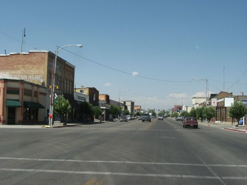 A view of a small town street with shops, trees, and a clear blue sky. Cars are parked along the road.