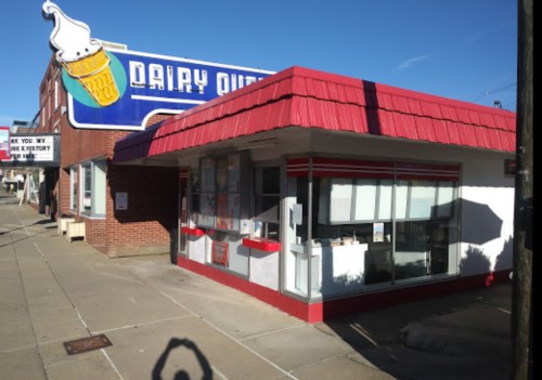 A small ice cream shop with a red roof and a large sign featuring an ice cream cone. Clear blue sky in the background.