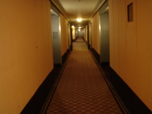 A dimly lit hotel corridor with patterned carpet and beige walls, leading to distant doors.