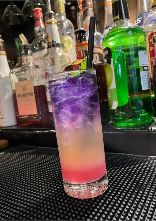 A colorful layered cocktail with purple, pink, and orange hues, garnished with mint, on a bar counter.
