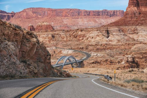A winding road leads to a modern bridge amidst red rock formations and desert landscape under a clear blue sky.