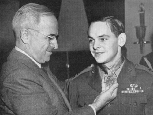 A man in a suit presents a medal to a young soldier in uniform, smiling at each other in a formal setting.