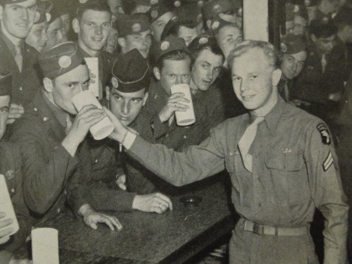 A group of soldiers in uniform enjoying drinks at a bar, with one soldier raising a glass in the foreground.