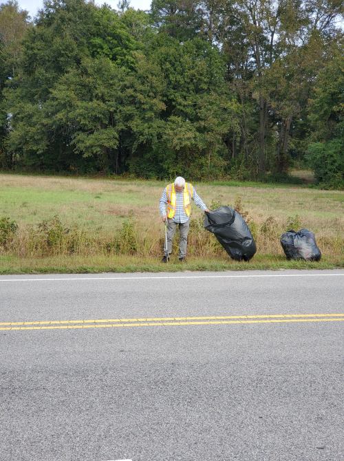 A person in a safety vest stands by the roadside, holding a trash bag near two other bags in a grassy area.