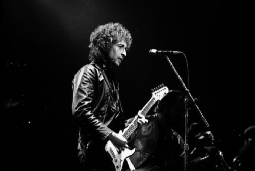 A musician in a leather jacket plays an electric guitar on stage, with a microphone in front of him.