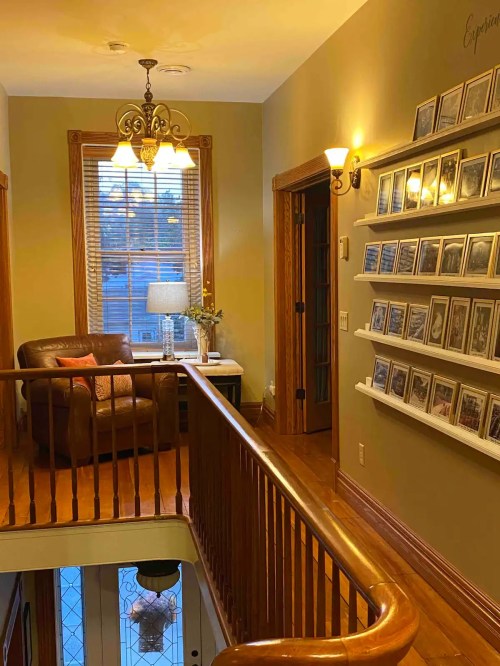 A cozy hallway with a brown armchair, a lamp, and a wall of framed photos, illuminated by warm lighting.