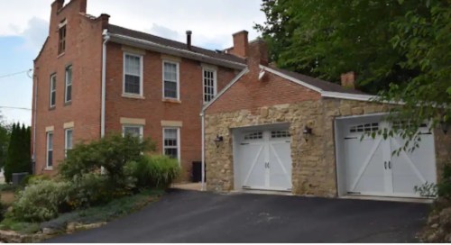 A two-story brick house with a stone garage, surrounded by greenery and a paved driveway.