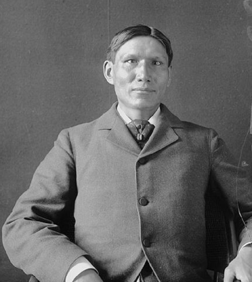 A seated man in a formal suit, with a neutral expression, against a plain background.